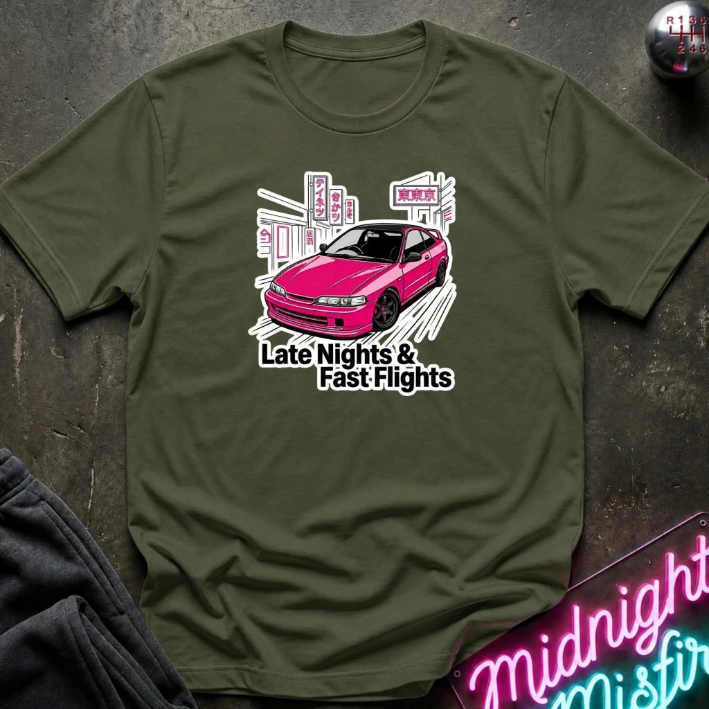 An olive green crewneck t-shirt laid flat on a dark textured surface. The shirt features a graphic print of a pink sports car in front of a city skyline with the text 'Late Nights & Fast Flights'.
