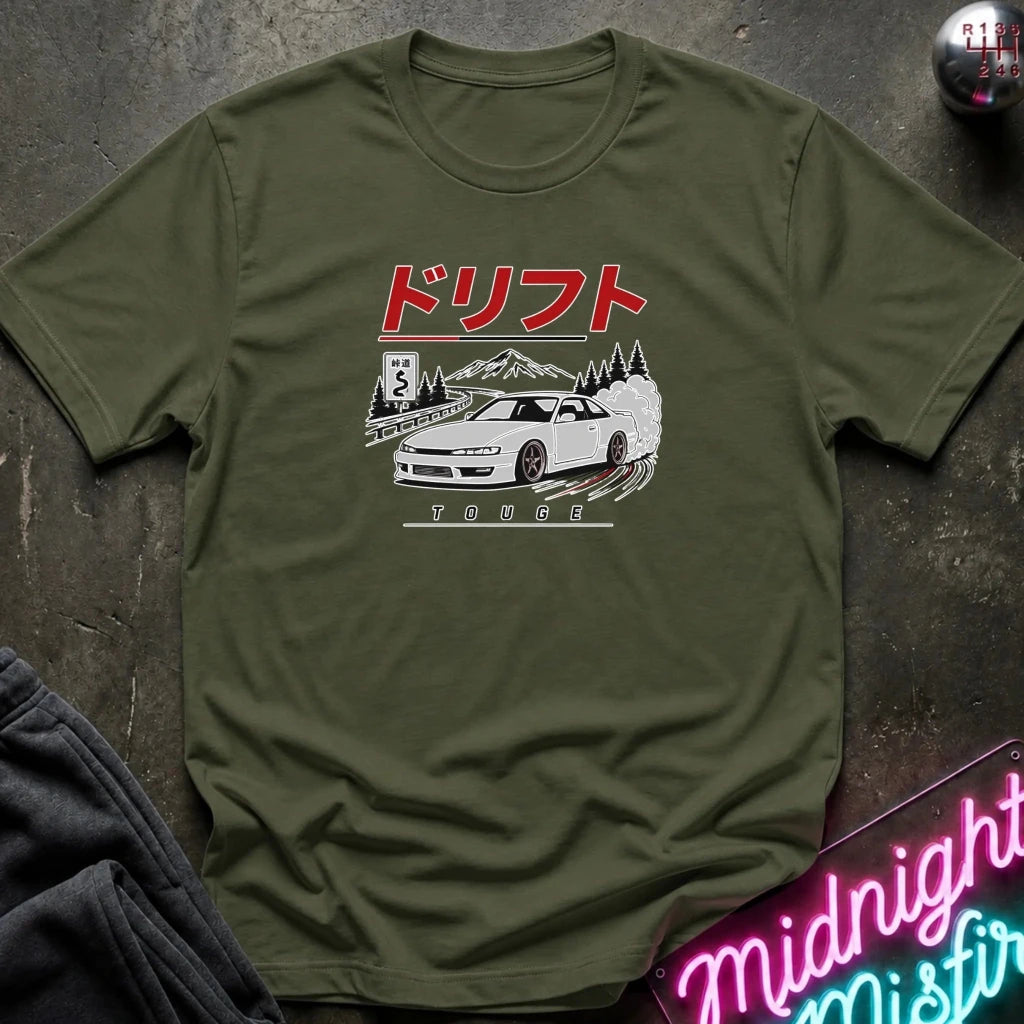 An olive green short-sleeve t-shirt with a Japanese drift car graphic, laid flat on a dark textured surface with a neon sign and dark pants partially visible.