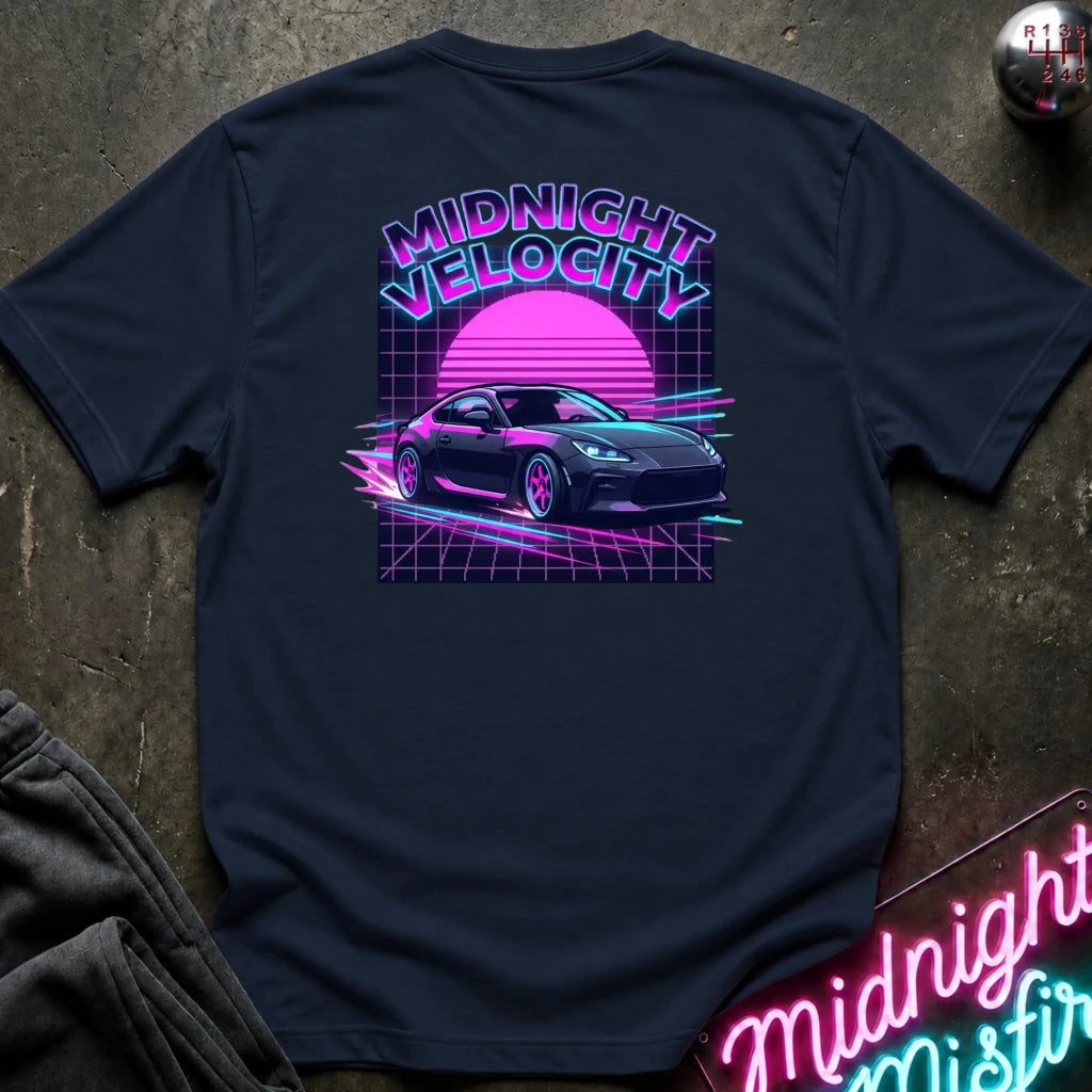 A navy blue short-sleeve t-shirt laid flat on a dark textured surface, showing the back graphic with the words 'MIDNIGHT VELOCITY' and a neon-style sports car illustration in pink, purple, and blue. 