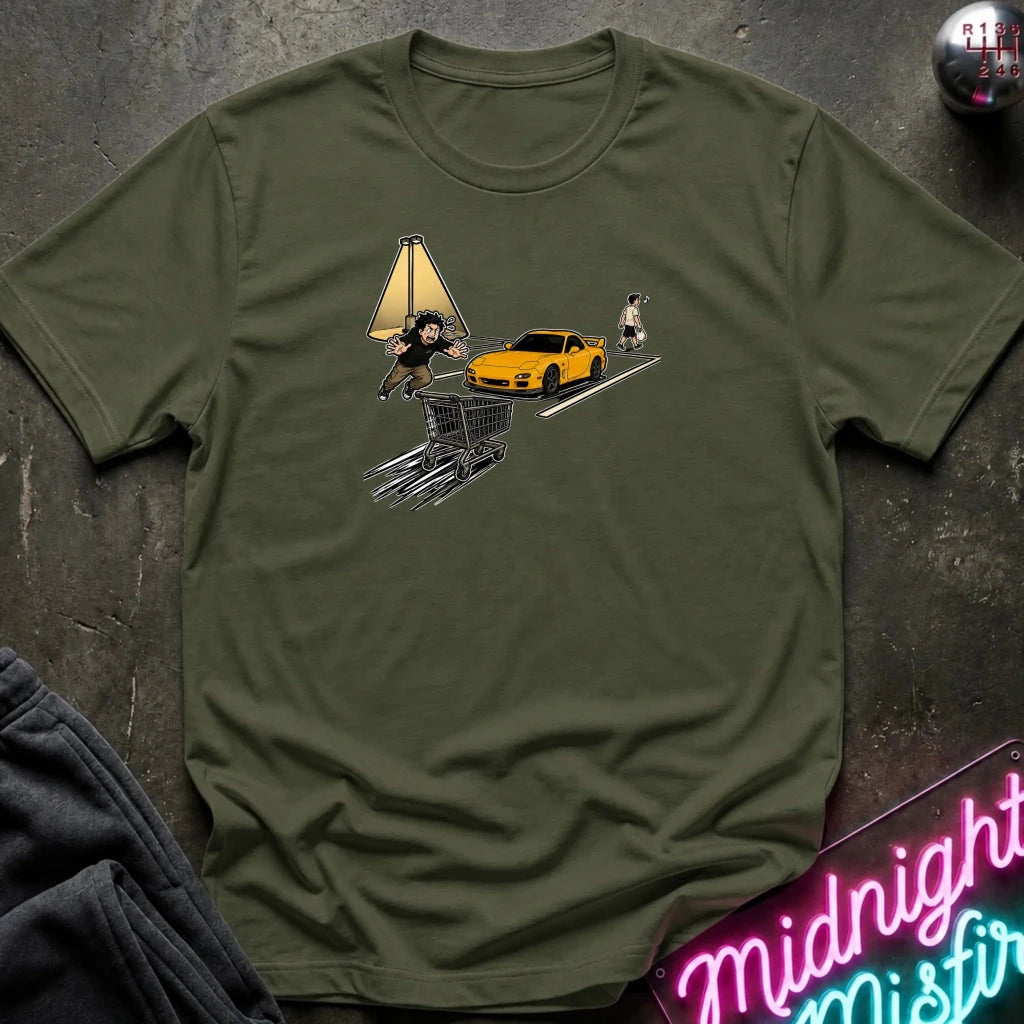 An olive green short-sleeve t-shirt laid flat on a dark textured surface, featuring a graphic print of a yellow sports car, a shopping cart, a dog, and a streetlamp. A neon sign and folded dark fabric are partially visible in the corners.