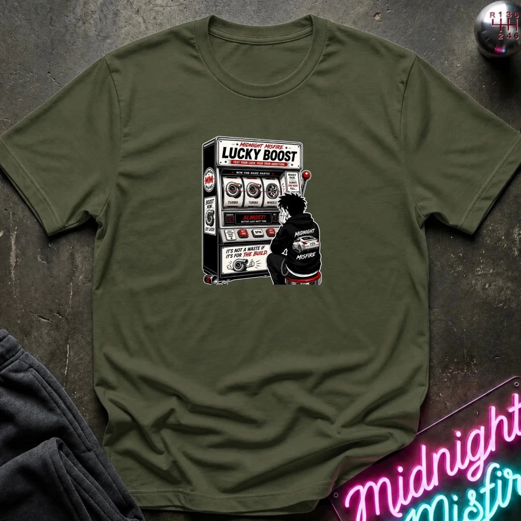 An olive green crewneck t-shirt laid flat on a dark textured surface, featuring a graphic print of a person sitting in front of a slot machine with the text 'LUCKY BOOST'.
