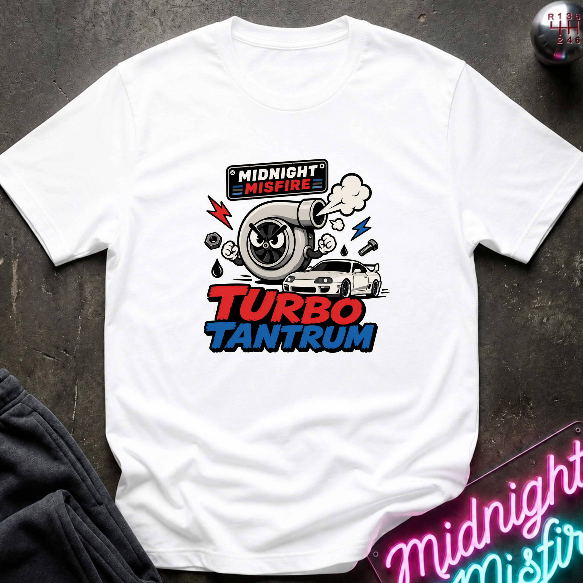 A white short-sleeve graphic T-shirt laid flat on a dark textured surface. The shirt features a colorful front print with a turbocharger, smoke, lightning bolts, a small car, and the text 'MIDNIGHT MISFIRE' and 'TURBO TANTRUM.