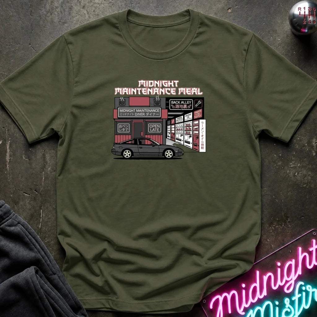 An olive green short-sleeve t-shirt laid flat on a dark textured surface, featuring a graphic print of a garage scene with the text 'MIDNIGHT MAINTENANCE MEAL' and a small car illustration. A neon sign and folded dark fabric are partially visible around it.