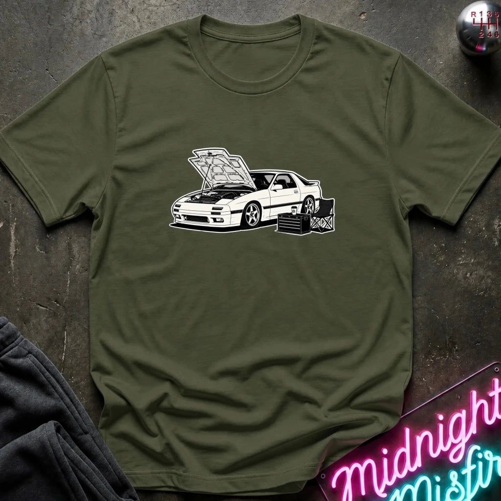 An olive green short-sleeve t-shirt with a white graphic of a sports car with its hood open, styled flat on a dark textured background with a neon sign and dark pants partially visible.