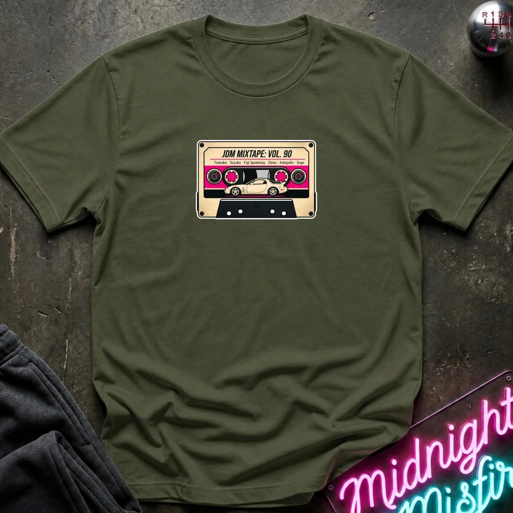 An olive green crewneck t-shirt laid flat on a dark textured surface, featuring a retro cassette tape graphic on the chest with the text 'JDM MIXTAPE VOL.