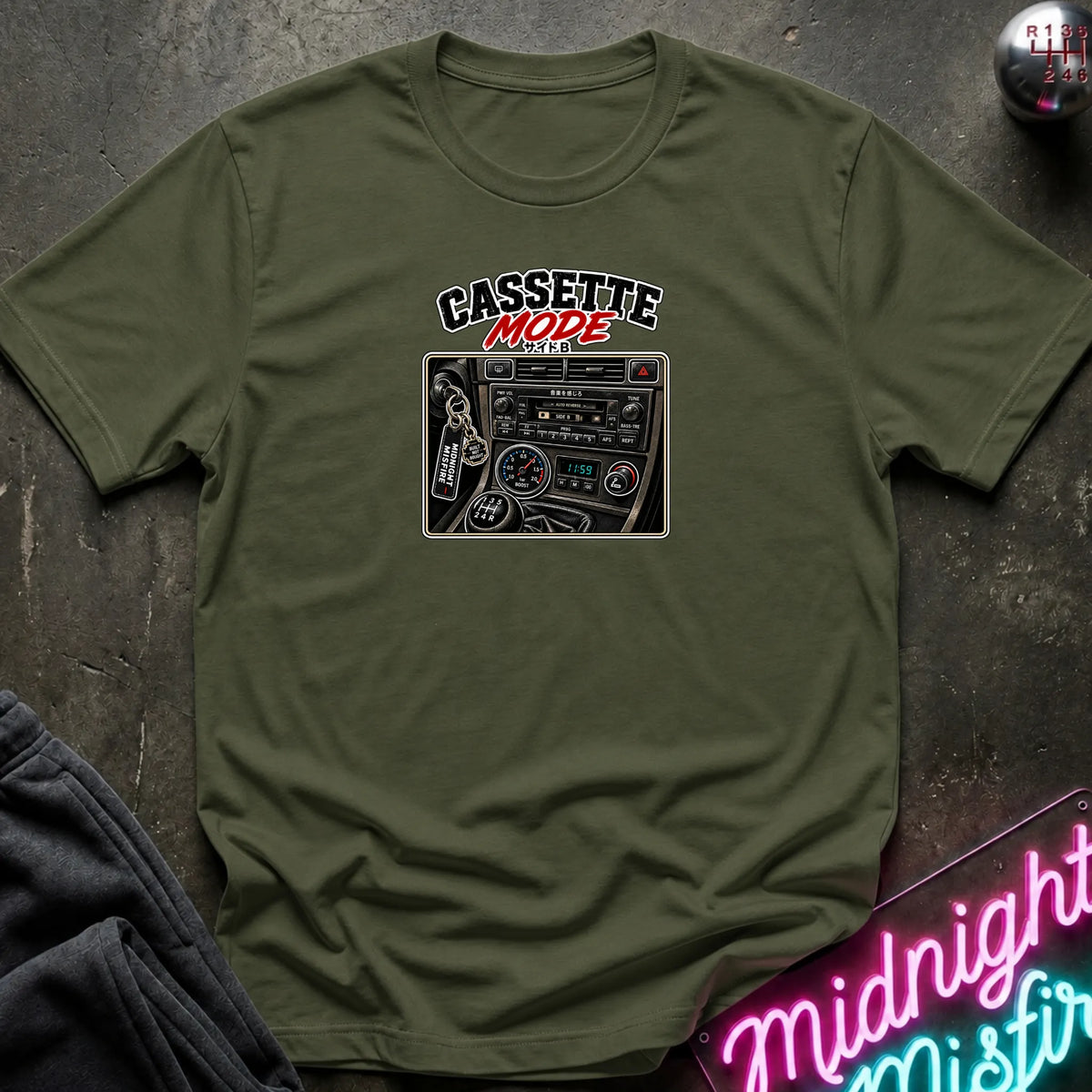 A olive short-sleeve t-shirt laid flat on a dark textured surface, featuring a graphic print of a cassette radio with the text 'CASSETTE MODE' in black and red. A neon sign and dark pants are partially visible as props.
