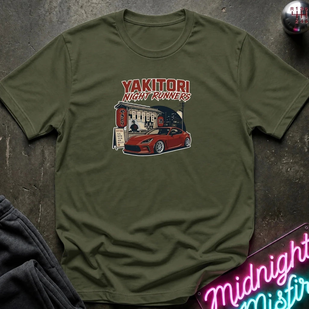 An olive green short-sleeve t-shirt laid flat on a dark textured surface, featuring a graphic print of a red sports car in front of a building with the text 'YAKITORI NIGHT RUNERS'.