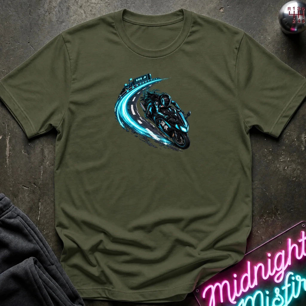 An olive green short-sleeve t-shirt with a graphic of a black motorcycle drifting on a blue-tinted road, laid flat on a dark textured surface with a neon sign and dark pants in the background.