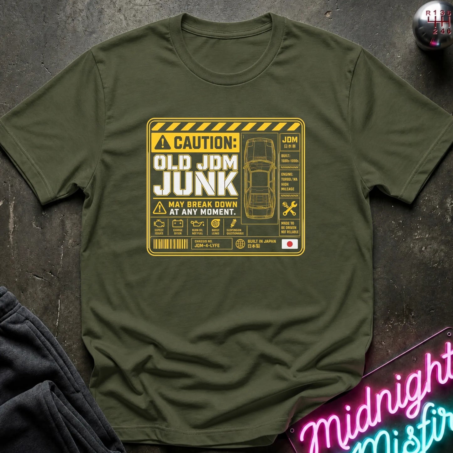 An olive green crewneck t-shirt featuring a yellow and black warning-style graphic that reads 'CAUTION: OLD JDM JUNK MAY BREAK DOWN AT ANY MOMENT.' with a car outline and technical-style icons, shown laid flat on a dark textured surface with partial props at the edges.