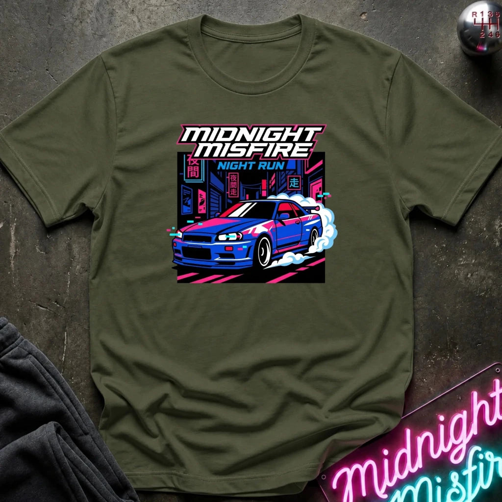 An olive green short-sleeve t-shirt with a graphic print of a blue and red sports car on a neon city street, with the text 'MIDNIGHT MISFIRE' and 'NIGHT RUN'. The shirt is laid flat on a dark textured surface with a neon sign and dark pants partially visible.