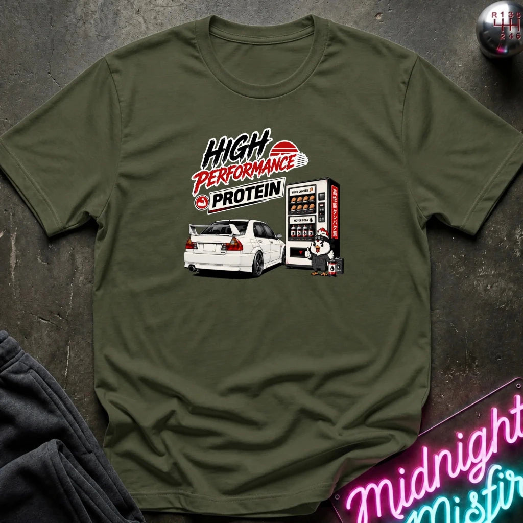 An olive green crewneck t-shirt laid flat on a dark textured surface. The shirt features a graphic print of a white sports car next to a vending machine with the text 'HIGH PERFORMANCE PROTEIN'.