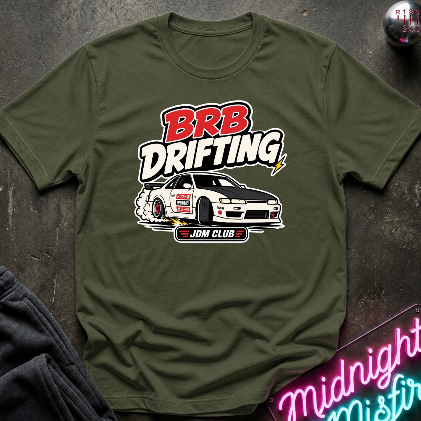 An olive green short-sleeve t-shirt with a 'BRB Drifting' graphic featuring a white drift car and 'JDM CLUB' text, laid flat on a dark textured surface with a neon sign and jeans partially visible.