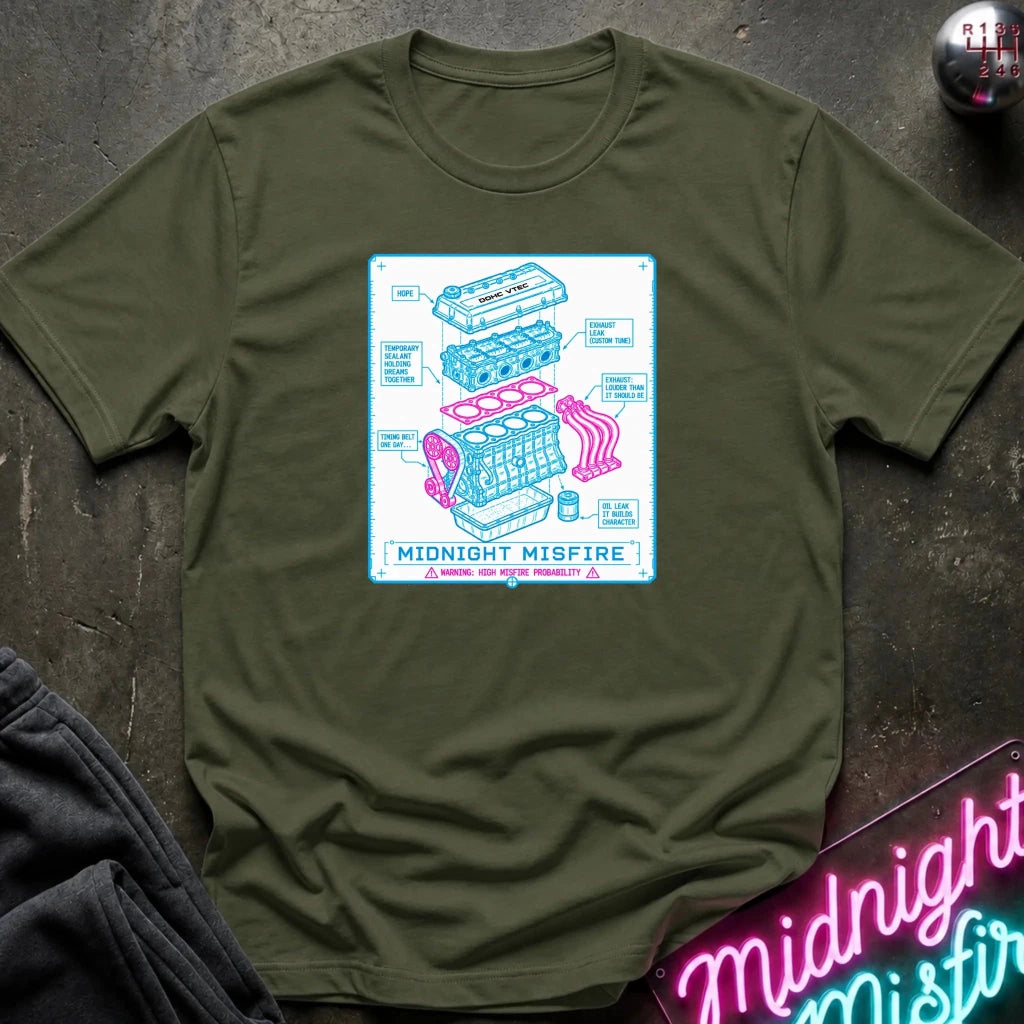 An olive green crewneck t-shirt with a colorful graphic print of a car engine labeled 'Midnight Misfire'. The shirt is laid flat on a dark textured surface with a neon sign and dark pants partially visible in the corners.