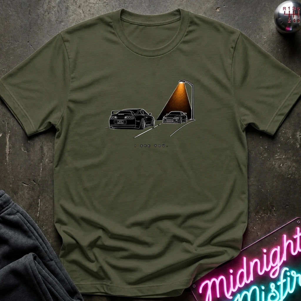 An olive green short-sleeve t-shirt laid flat on a dark textured surface, featuring a graphic print of two black sports cars passing each other.