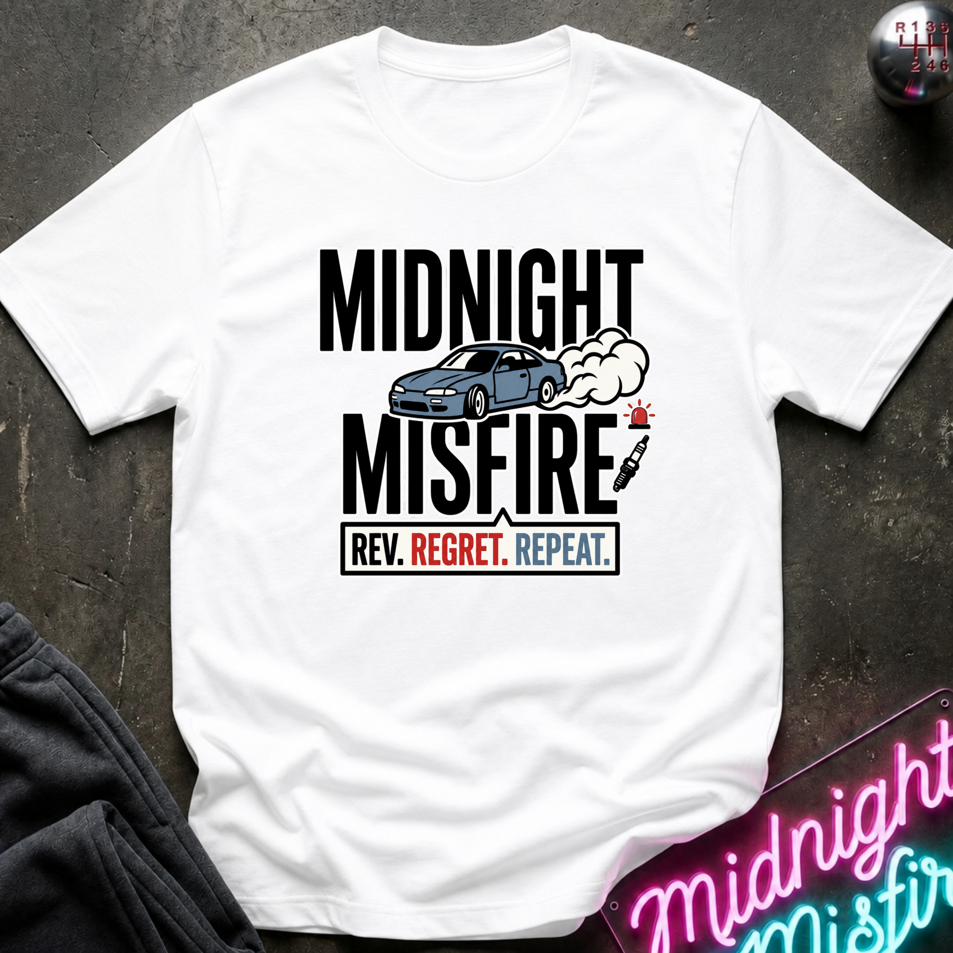 A white short-sleeve graphic T-shirt laid flat on a dark textured surface. The shirt features a black text design reading 'MIDNIGHT MISFIRE' with a blue car, smoke, and a red/white/black tag below.