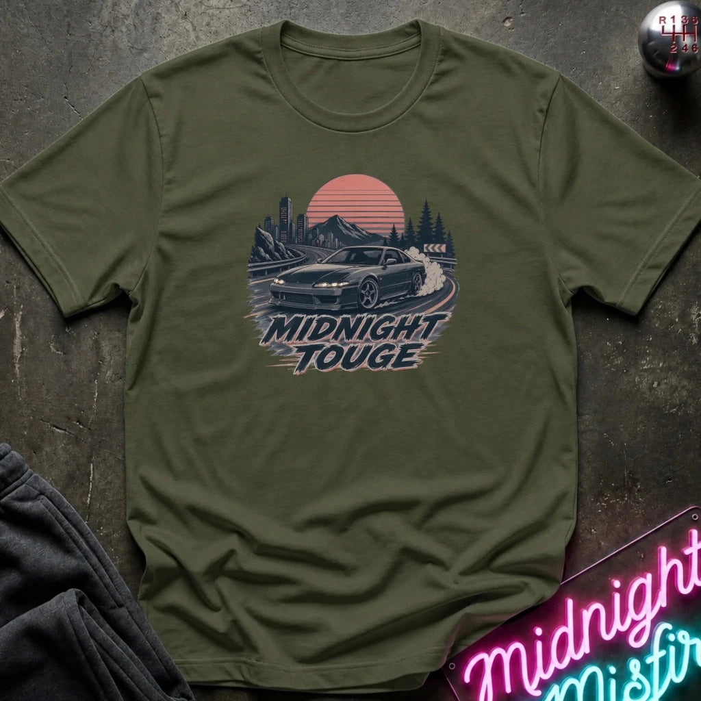 An olive green short-sleeve t-shirt laid flat on a dark textured surface, featuring a retro-style graphic of a car drifting around a track with mountains and a sunset, and the text 'MIDNIGHT TOUGHE' printed below. A neon sign and folded dark fabric are partially visible around it.