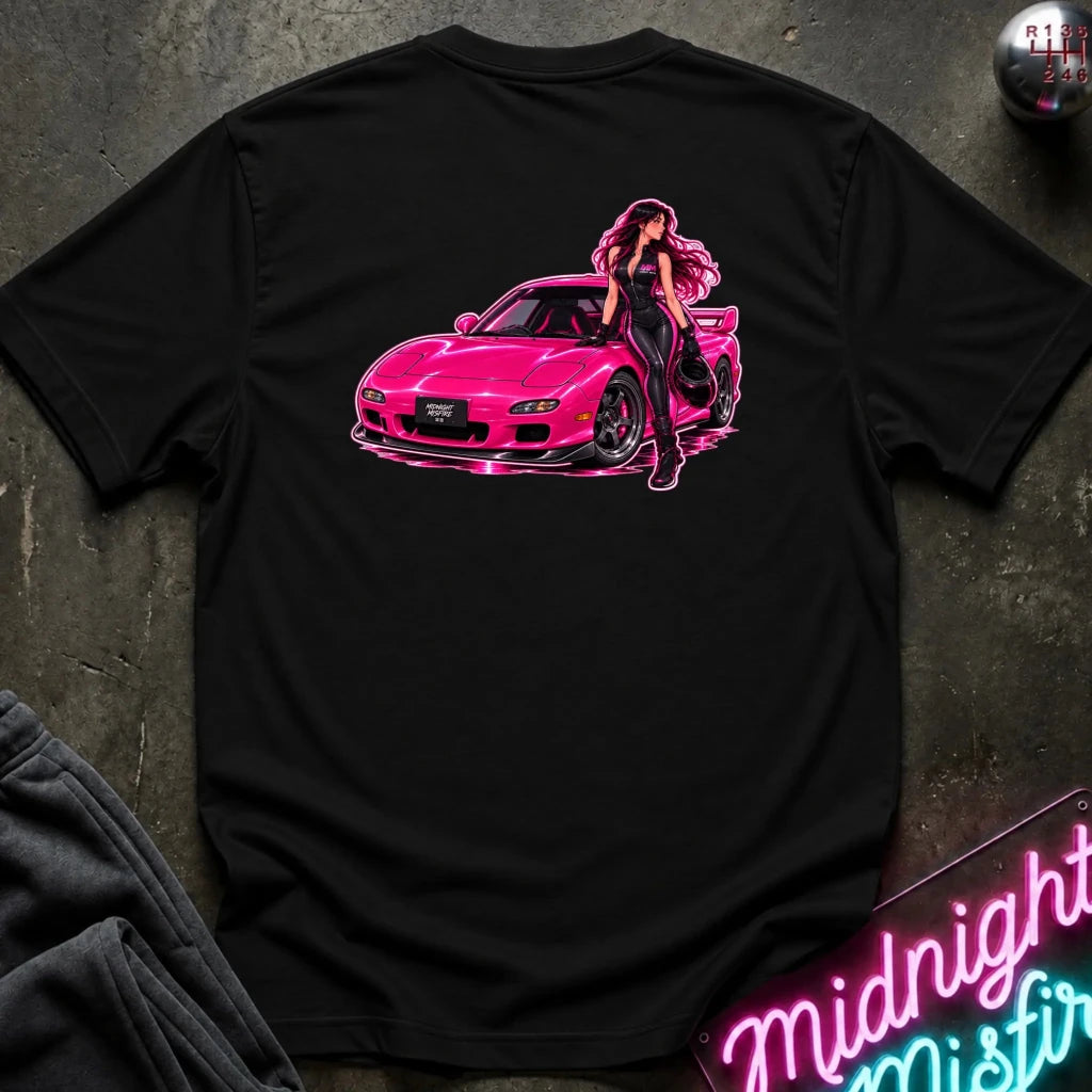 A black short-sleeve t-shirt laid flat on a dark textured surface, featuring a large graphic print of a pink sports car with a woman leaning against it. The shirt is styled with dark gray sweatpants, a decorative ball, and a neon sign in the background.