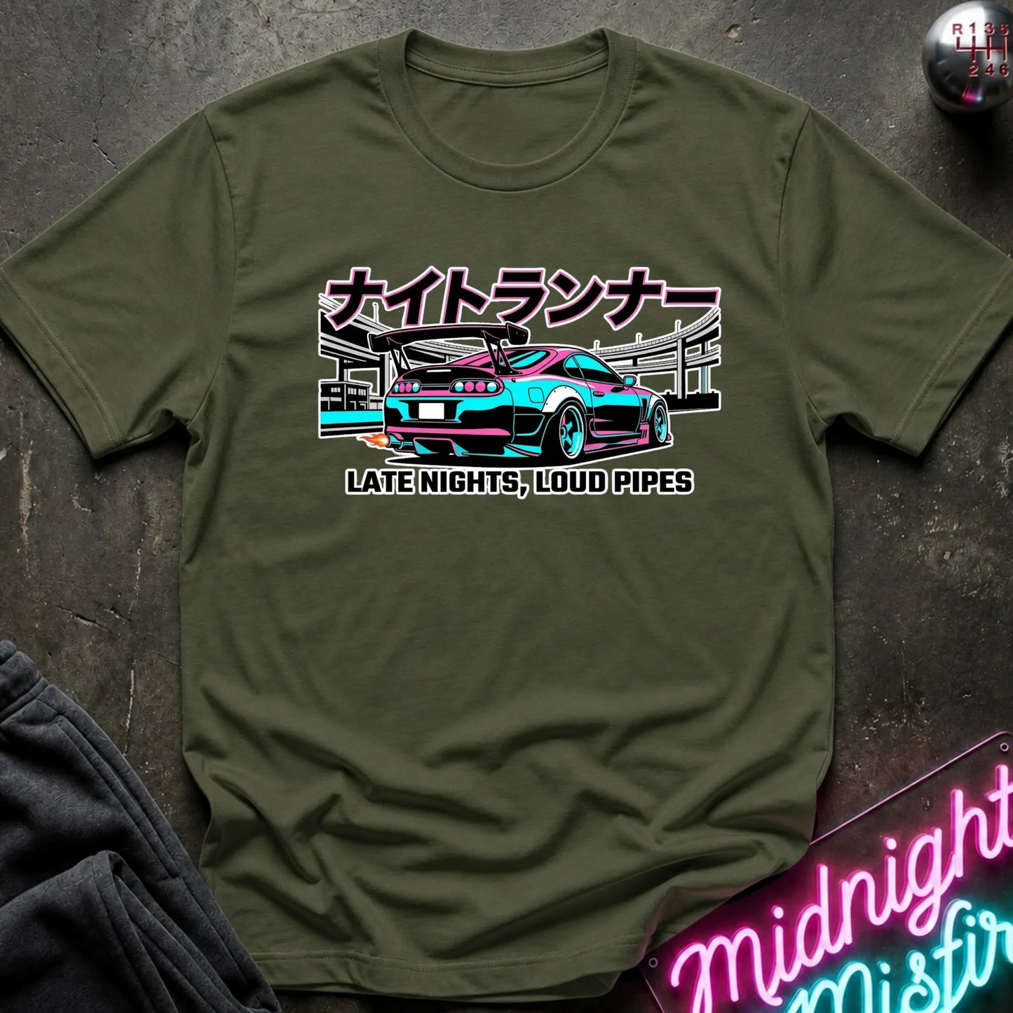 An olive green graphic t-shirt featuring a stylized cyan and pink sports car with Japanese text and 'LATE NIGHTS, LOUD PIPES' underneath, laid flat on a dark textured surface with a neon sign and dark pants partially visible.