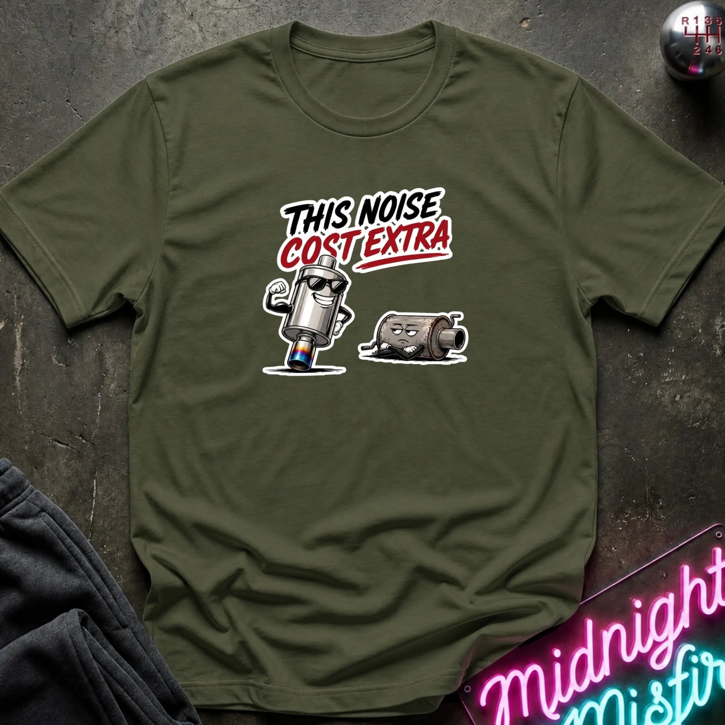 An olive green short-sleeve t-shirt laid flat on a dark textured surface. The shirt features a graphic print of a cartoon character with the text 'THIS NOISE COST EXTRA' in white and red.