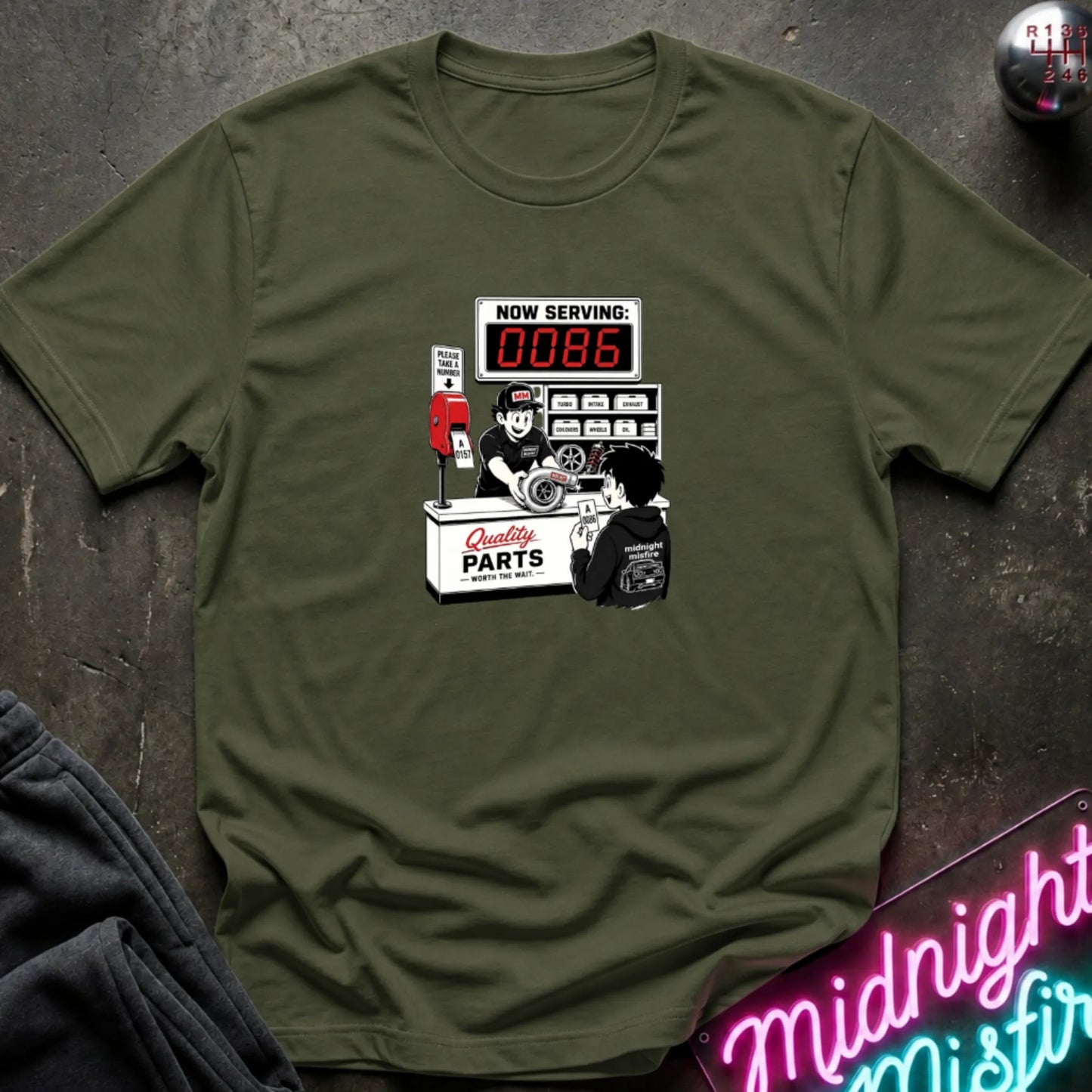 An olive green   short-sleeve t-shirt with a retro-style graphic of a garage attendant at a 'Quality Parts' counter, displayed flat on a dark textured surface with a neon sign and sweatpants partially visible.