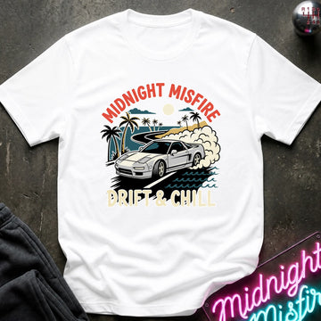 A white short-sleeve graphic T-shirt laid flat on a dark textured surface. The shirt features a colorful front print with the words 'MIDNIGHT MISFIRE' and 'DRIFT & CHILL' around an illustration of a silver sports car on a beach with palm trees and waves.