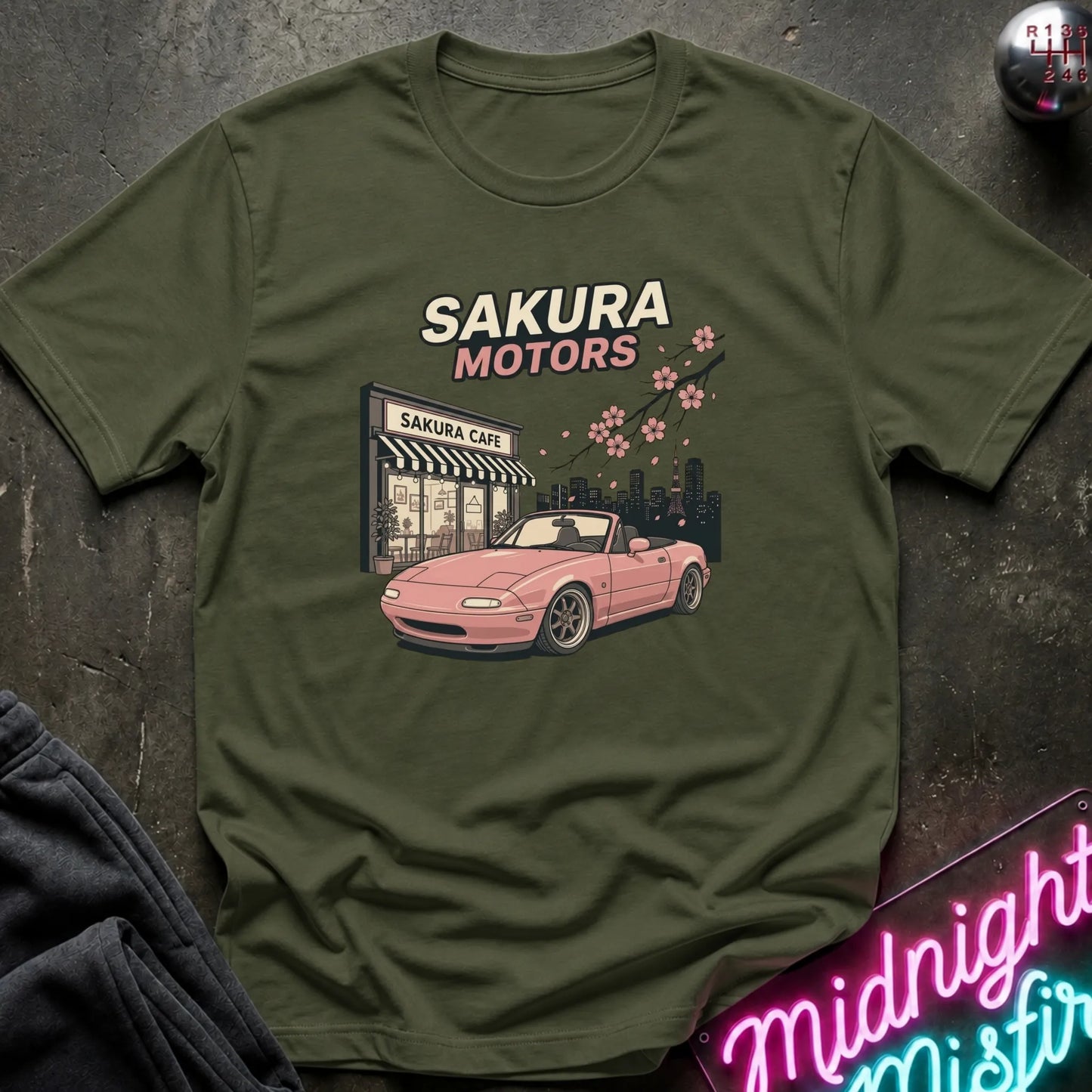 An olive green graphic t-shirt laid flat on a dark textured surface. The shirt features a graphic print of a pink convertible car in front of a 'Sakura CAFE' with cherry blossoms and the text 'SAKURA MOTORS'.