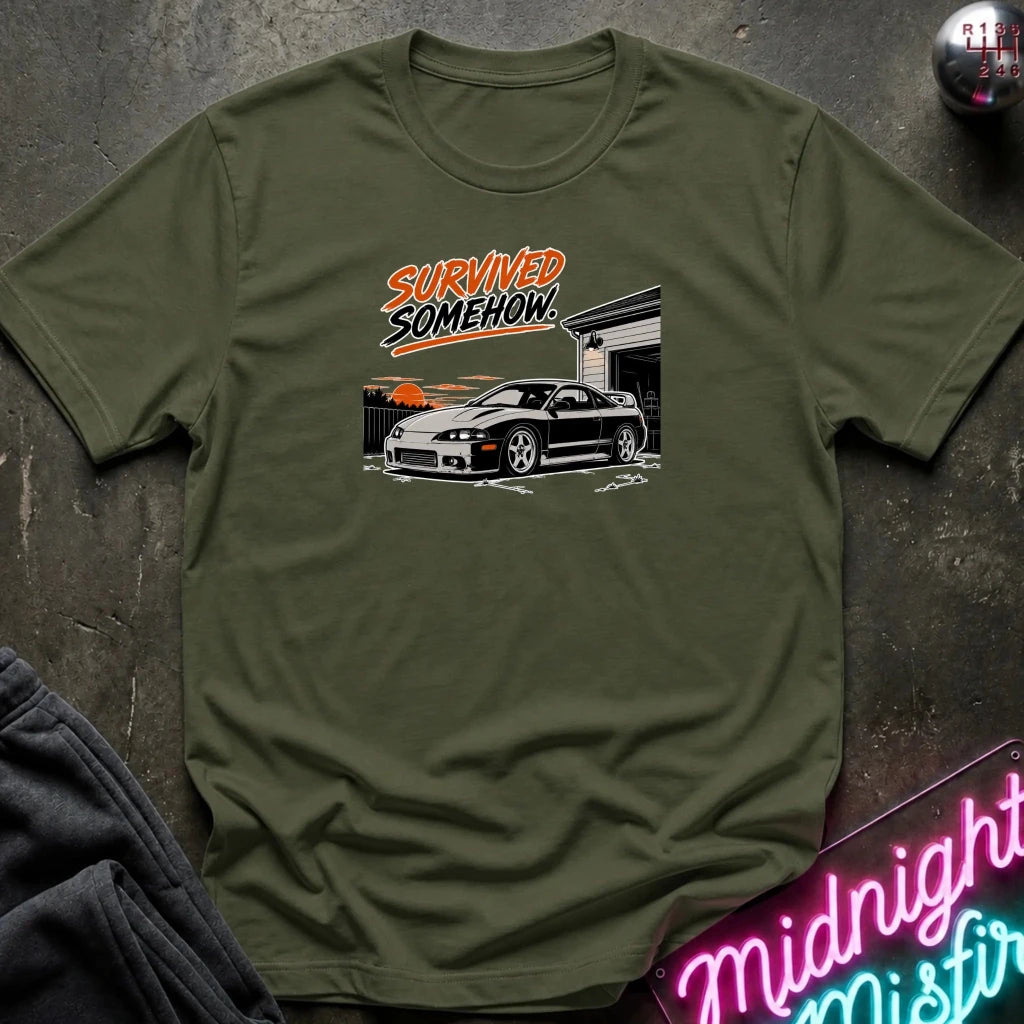 An olive green short-sleeve t-shirt with a graphic print of a black sports car and the text 'SURVIVED SOMEHOW' in orange and black, laid flat on a dark textured surface.