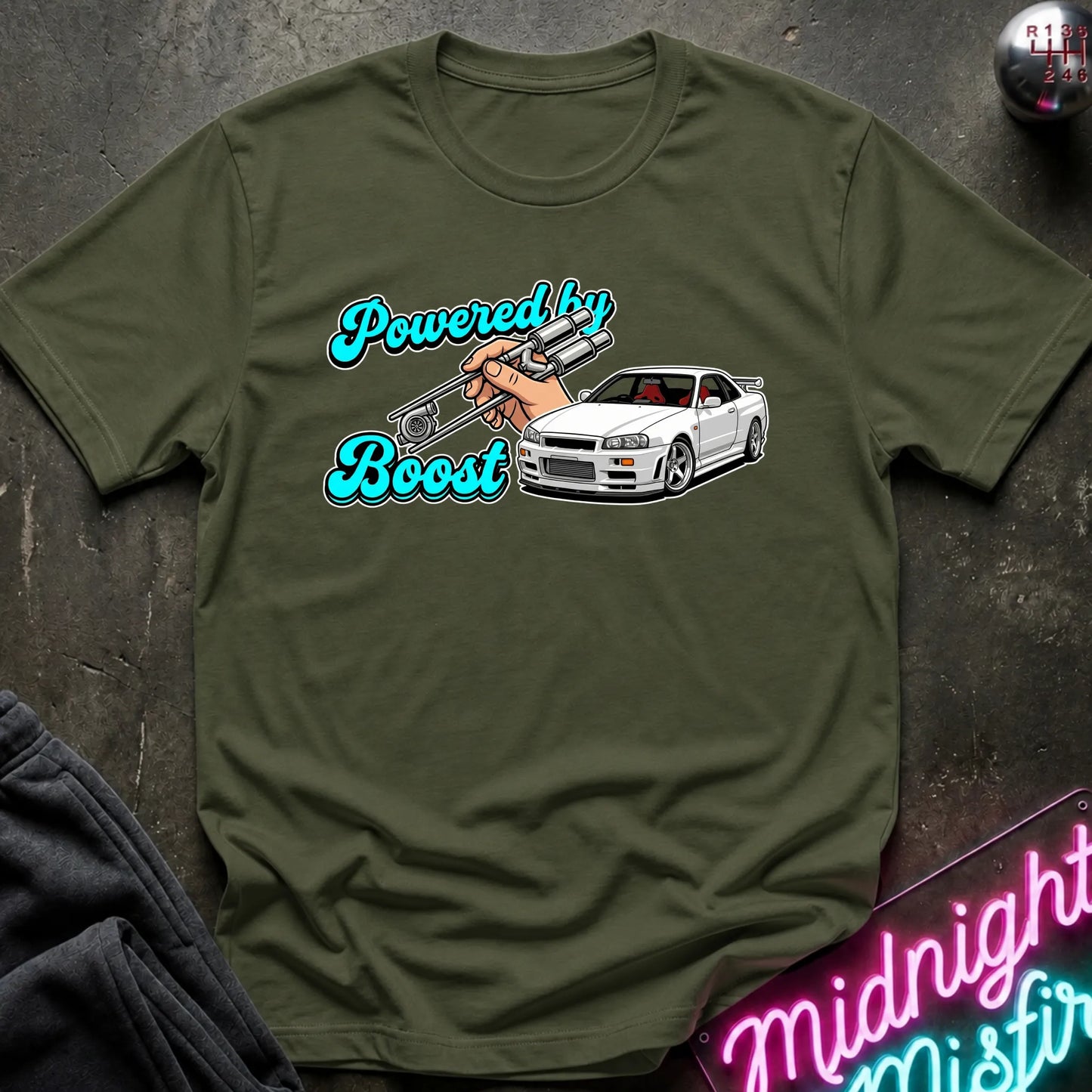 An olive green graphic t-shirt laid flat on a dark textured surface. The graphic features the text 'Powered by Boost' in a teal cursive font, with a hand holding a silver turbocharger, alongside a white sports car.