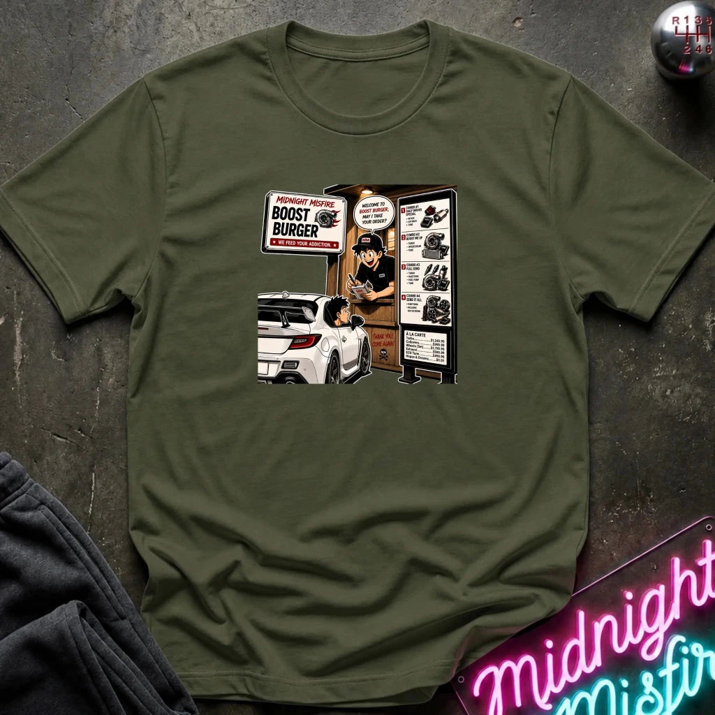 An olive green crewneck t-shirt with a retro-style graphic print of a fast-food burger joint advertisement, laid flat on a dark textured surface with a neon sign and dark fabric in the foreground.
