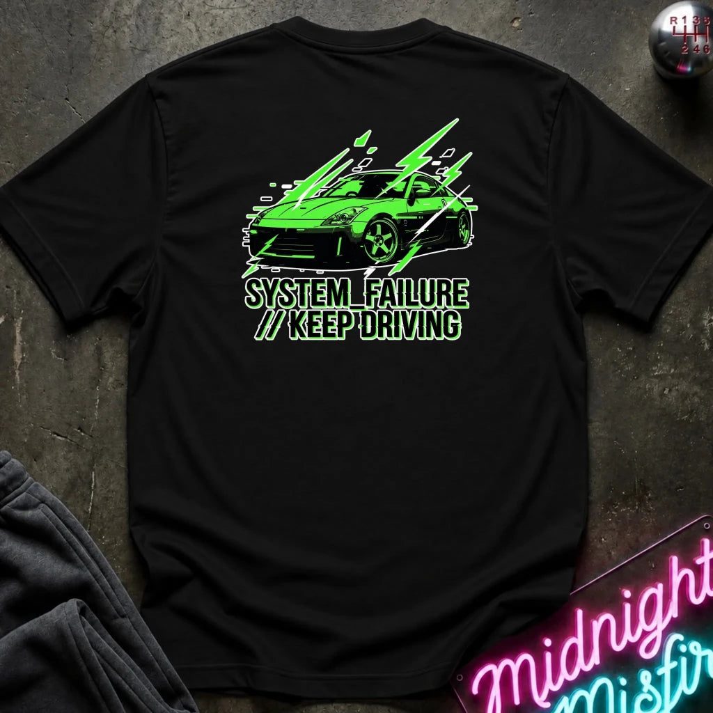 black short-sleeve t-shirt laid flat on a dark textured surface, featuring a graphic of a green sports car with text 'SYSTEM FAILURE // KEEP DRIVING' in green. A neon sign and dark clothing are partially visible as props.