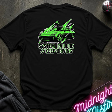 black short-sleeve t-shirt laid flat on a dark textured surface, featuring a graphic of a green sports car with text 'SYSTEM FAILURE // KEEP DRIVING' in green. A neon sign and dark clothing are partially visible as props.