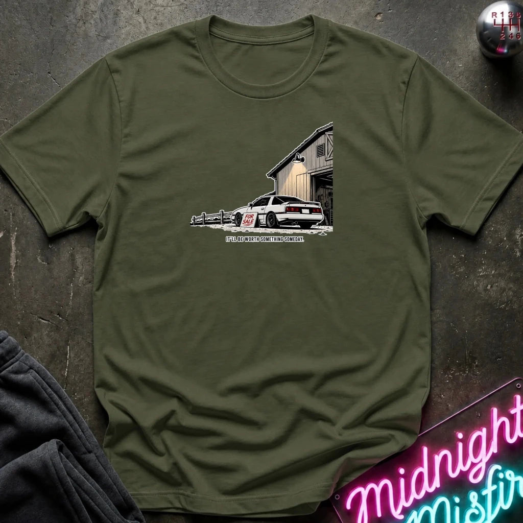 An olive green crewneck t-shirt featuring a graphic print of a white muscle car parked in front of a barn, styled flat on a dark textured surface with a neon sign and dark pants nearby.