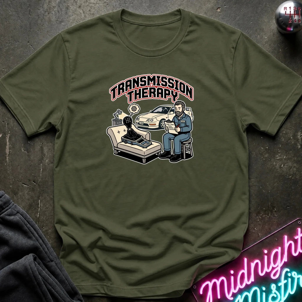 An olive green graphic t-shirt laid flat on a dark textured surface. The graphic features the words 'TRANSMISSION THERAPY' in arched pink letters above an illustration of a therapist sitting next to a patient in a chair, with a police car in the background.