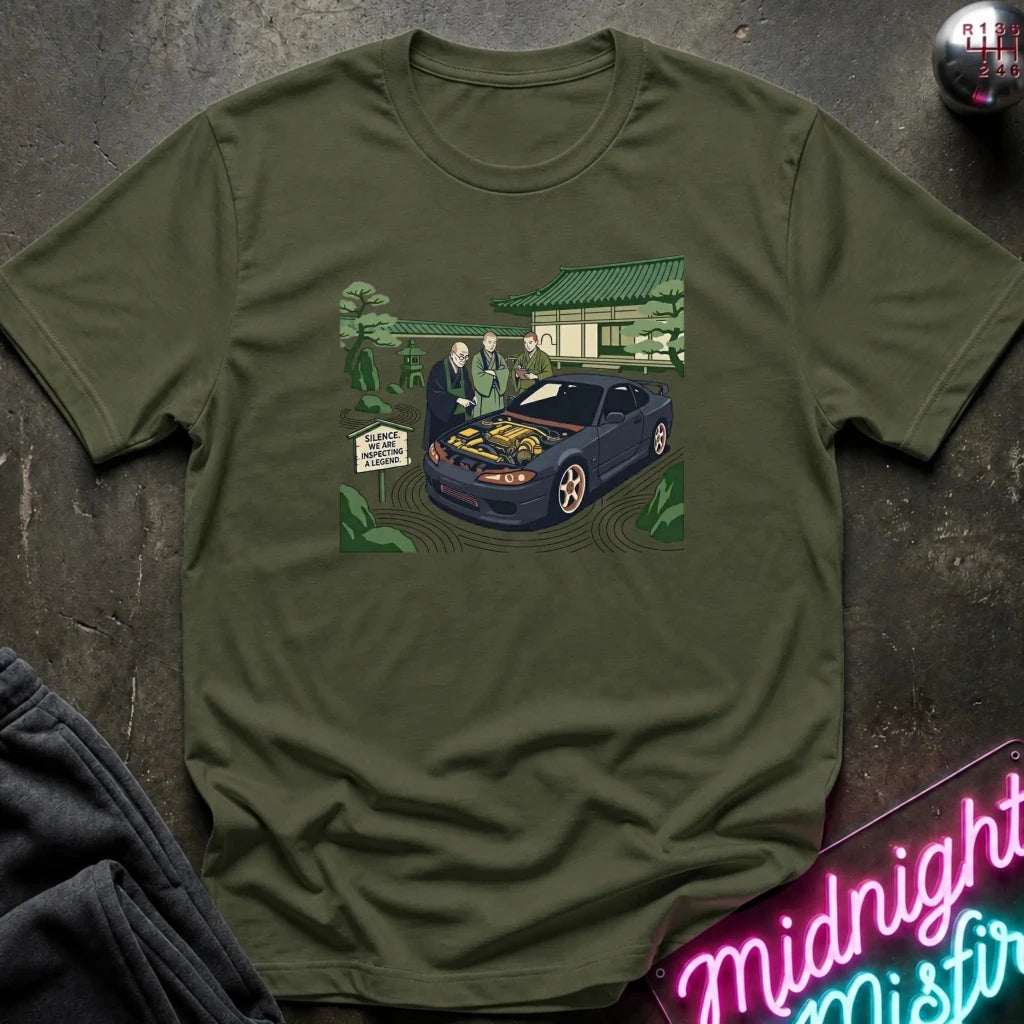 An olive green crewneck t-shirt laid flat on a dark textured surface, featuring a colorful graphic print of a blue sports car with its hood open, surrounded by characters in a garden setting. A neon sign and dark clothing are partially visible in the corners.