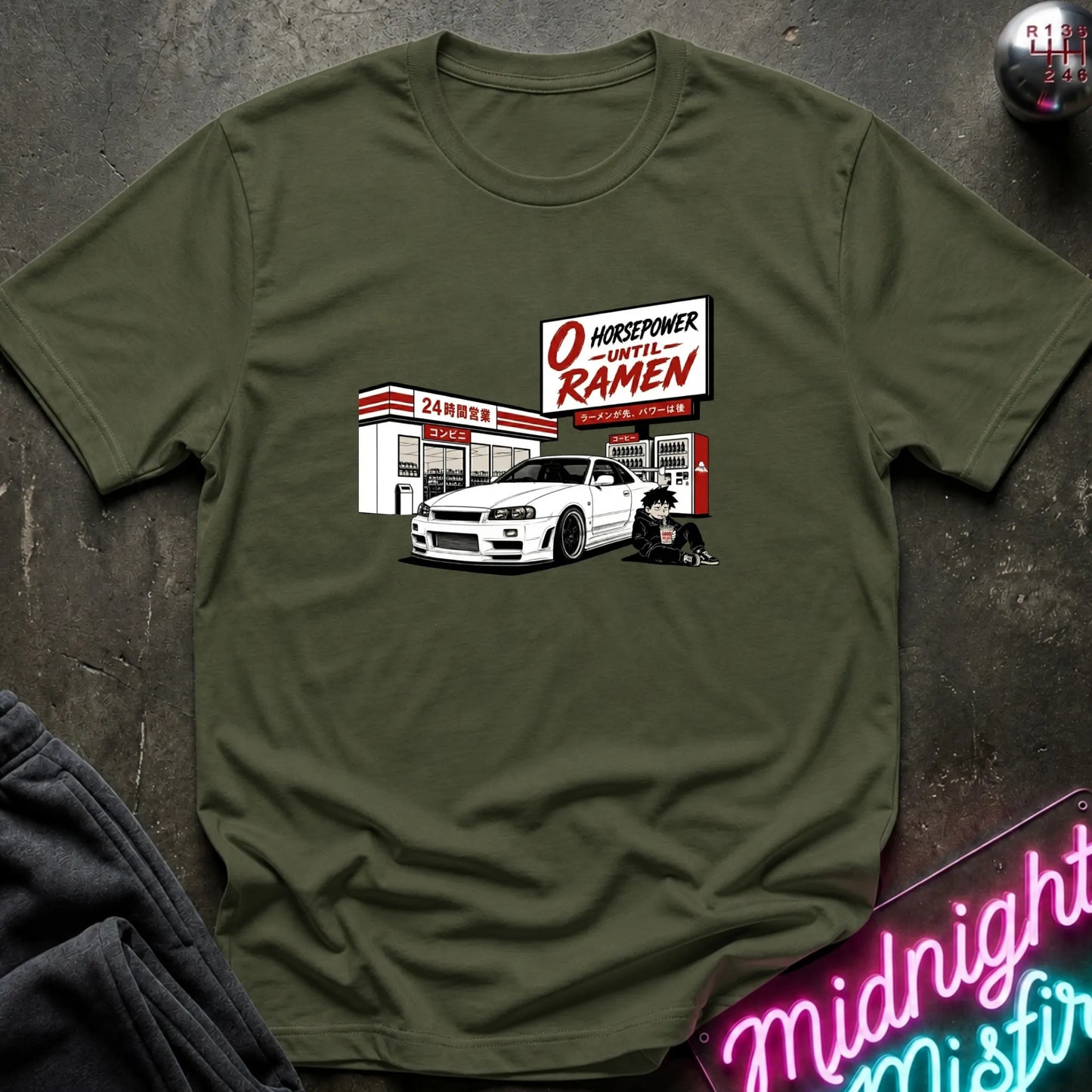 An olive green crewneck t-shirt laid flat on a dark textured surface, featuring a graphic print of a white sports car parked in front of a store with a sign that reads 'O HONORABLE -UNTIL RAMEN'.