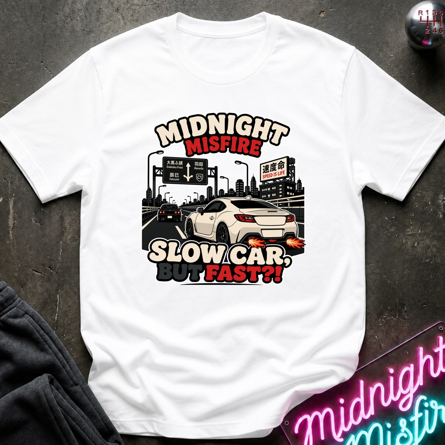 A white short-sleeve graphic T-shirt laid flat on a dark textured surface. The shirt features a printed design of a white sports car on a highway at night with the text 'MIDNIGHT MISFIRE' and 'SLOW CAR, STOP FAST?!' in red and black.