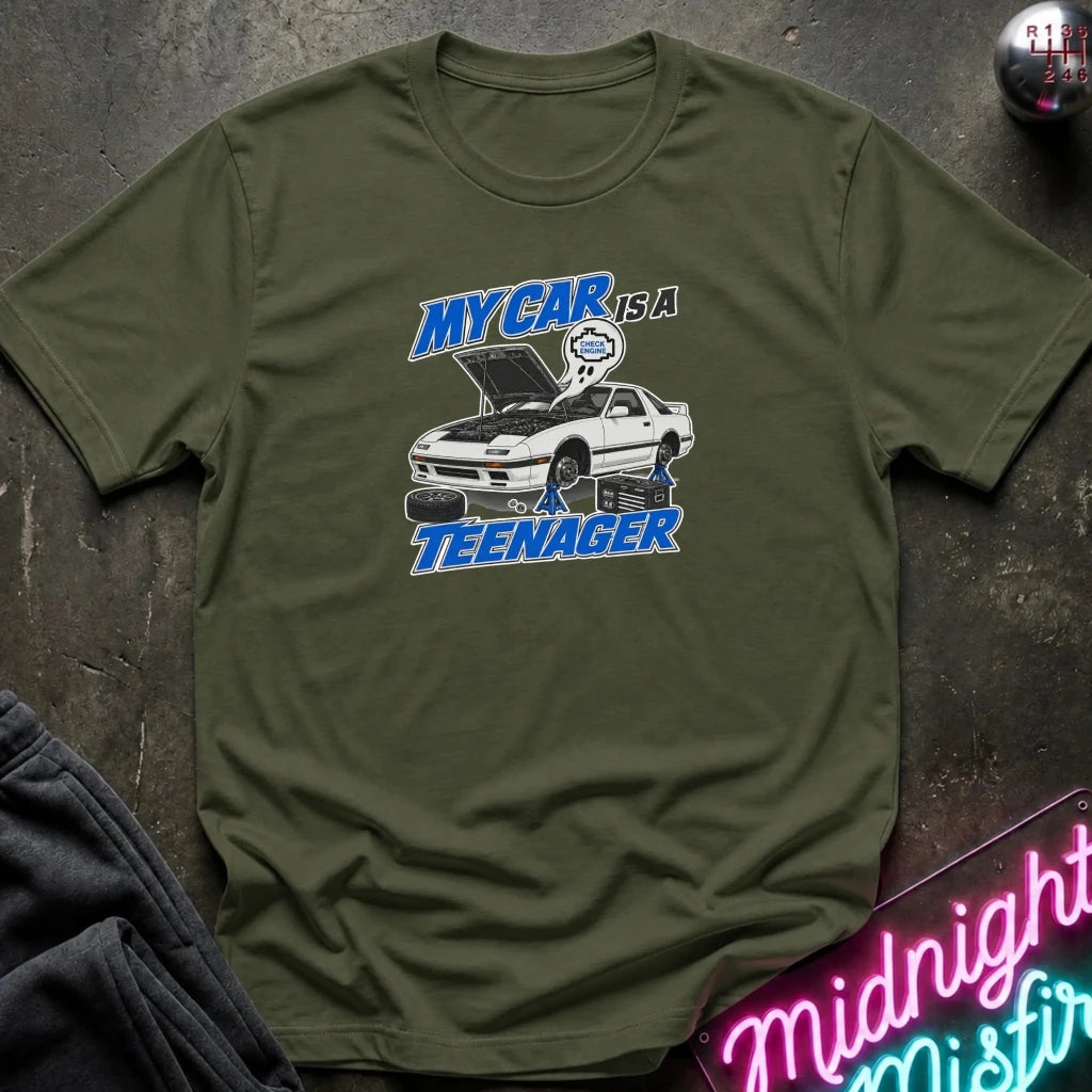 An olive green short-sleeve t-shirt with a graphic print of a white sports car and the text 'MY CAR IS A TEENAGER' in blue and white, laid flat on a dark textured surface.