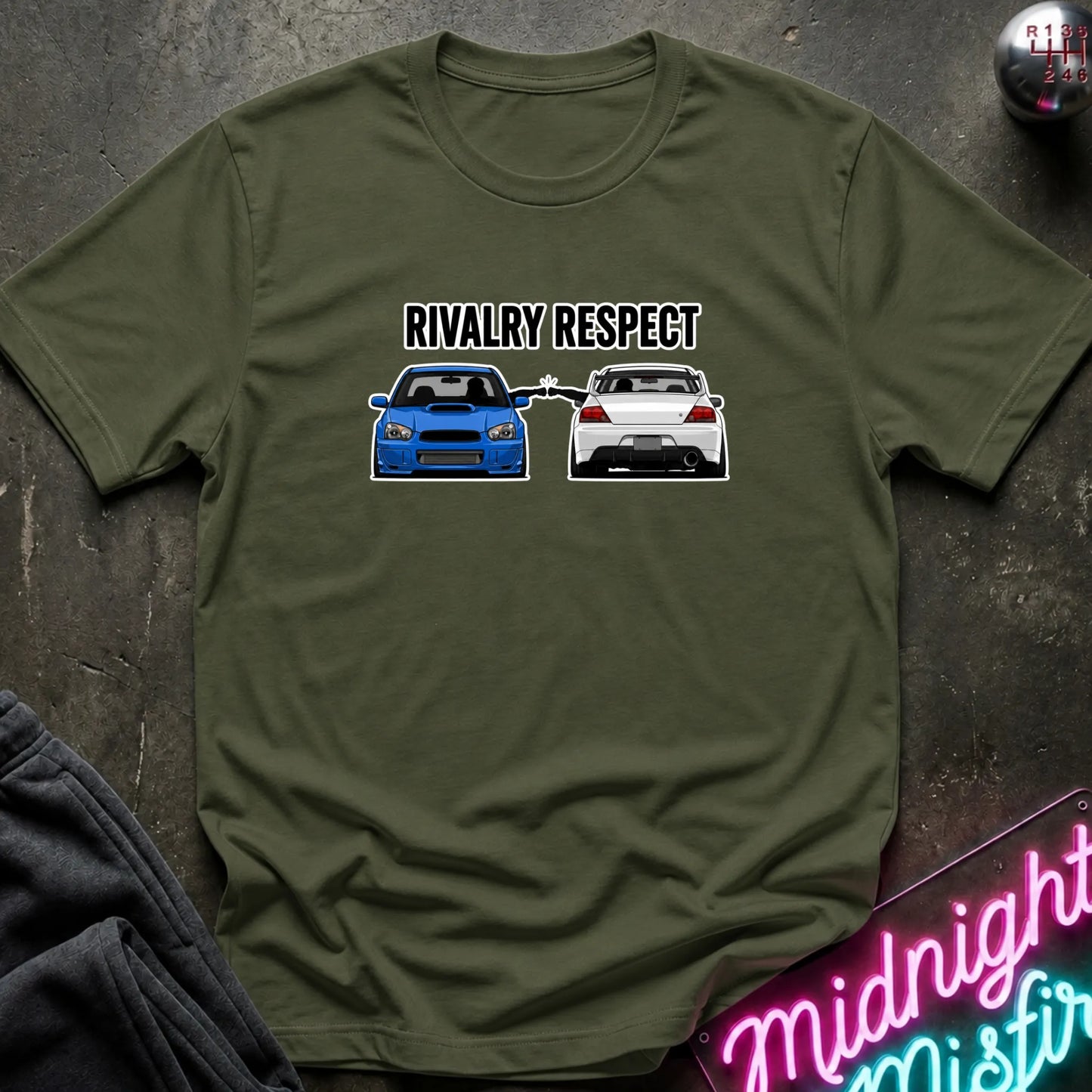 An olive green short-sleeve graphic t-shirt laid flat on a dark textured surface. The graphic features the text 'RIVALRY RESPECT' above a blue and white car, 