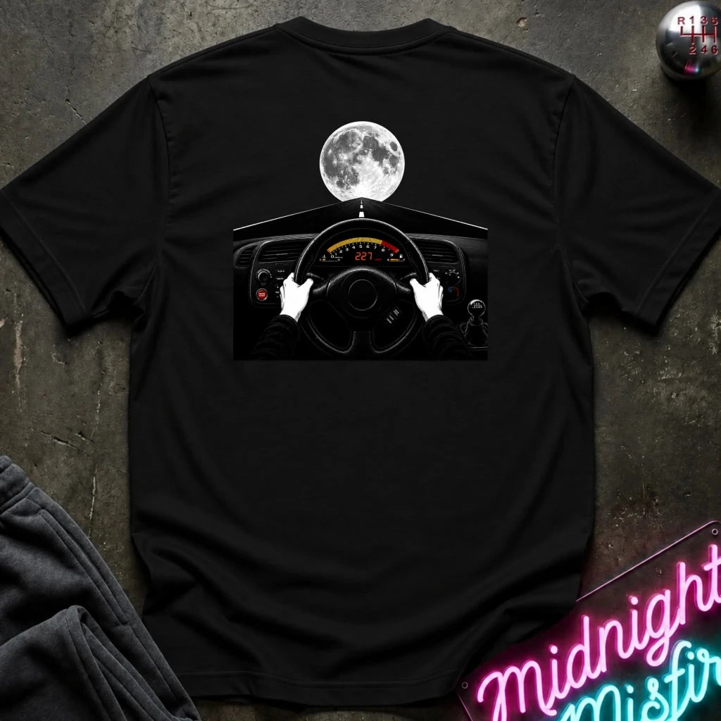 A black short-sleeve t-shirt laid flat on a dark textured surface, featuring a graphic print of a car dashboard and steering wheel with white hands and a full moon above. 