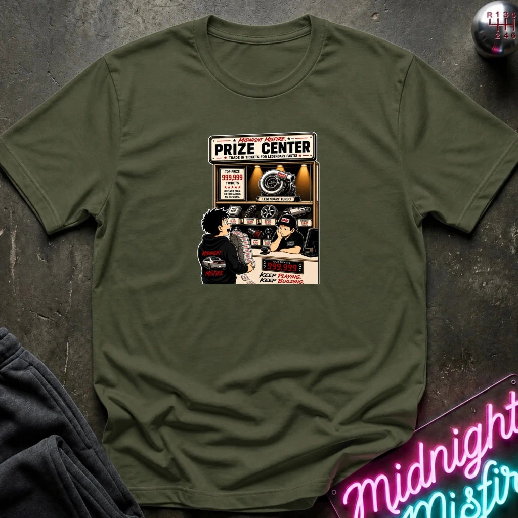 An olive green short-sleeve t-shirt with a vintage-style 'Prize Center' comic graphic on the chest, laid flat on a dark textured surface with a neon sign and dark pants partially visible.