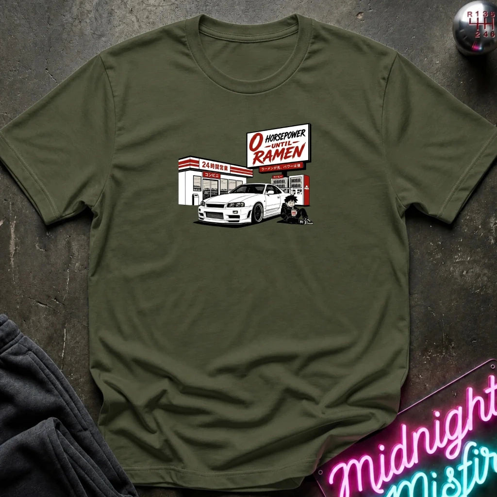 An olive green crewneck t-shirt laid flat on a dark textured surface, featuring a graphic print of a white sports car parked in front of a konbini.