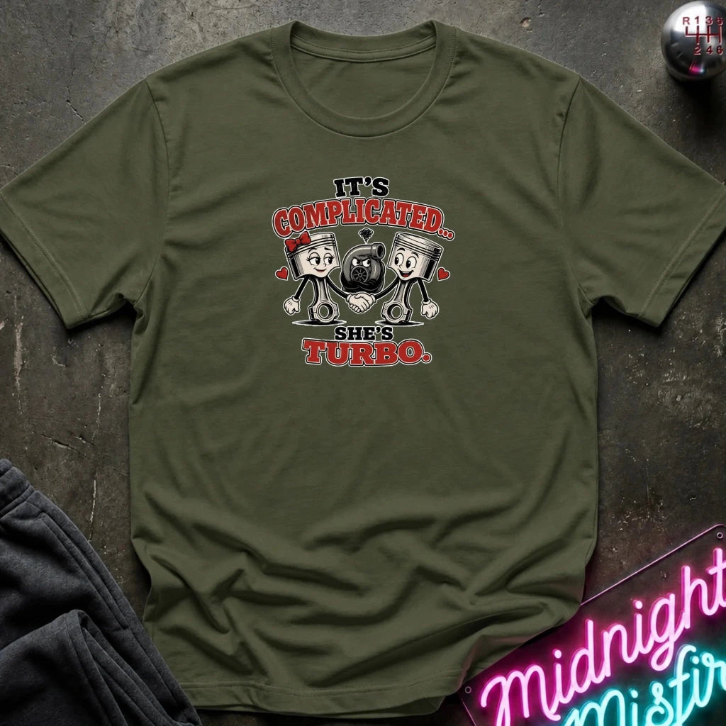 An olive green short-sleeve t-shirt with a cartoon graphic of two pistons holding a heart, with the text 'IT'S COMPLICATED. SHE'S TURBO.