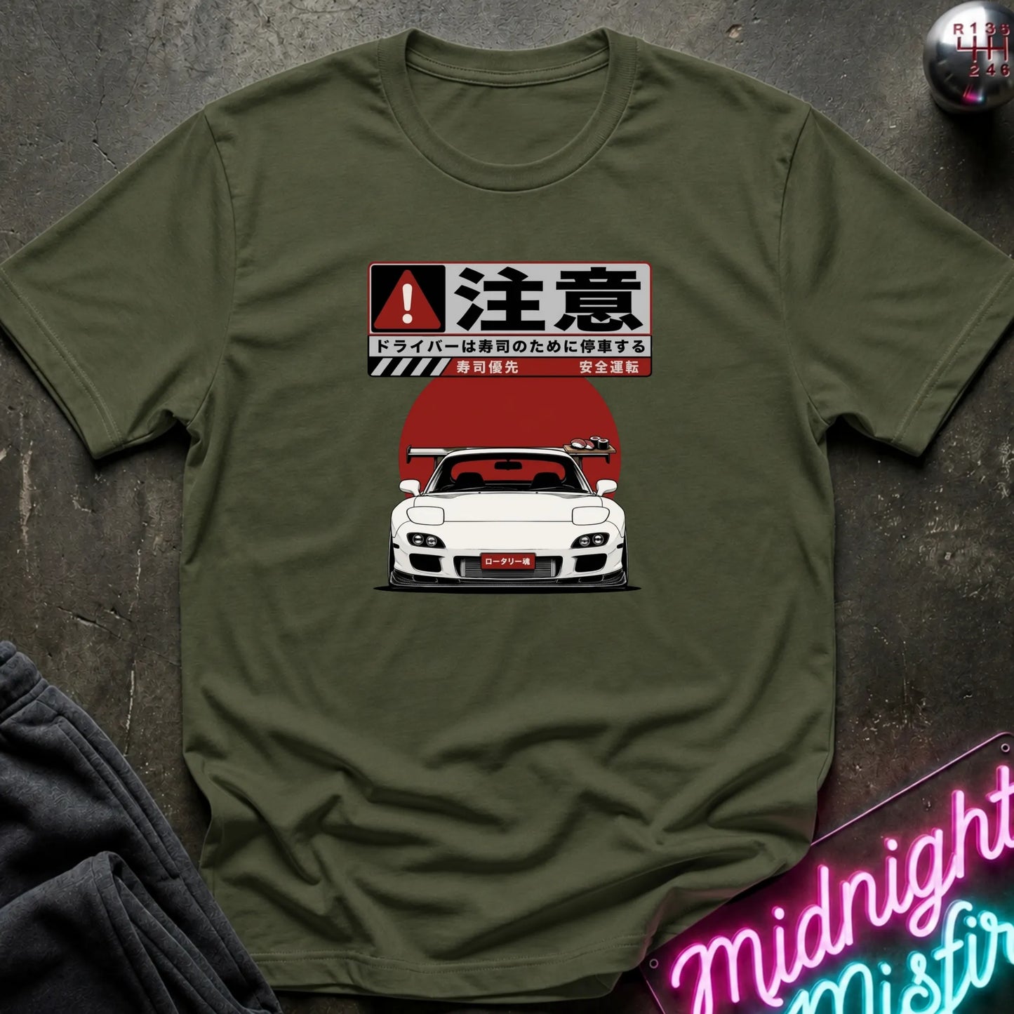 An olive green short-sleeve t-shirt with a Japanese warning sign and a white sports car graphic on the chest, laid flat on a dark textured surface with decorative props.
