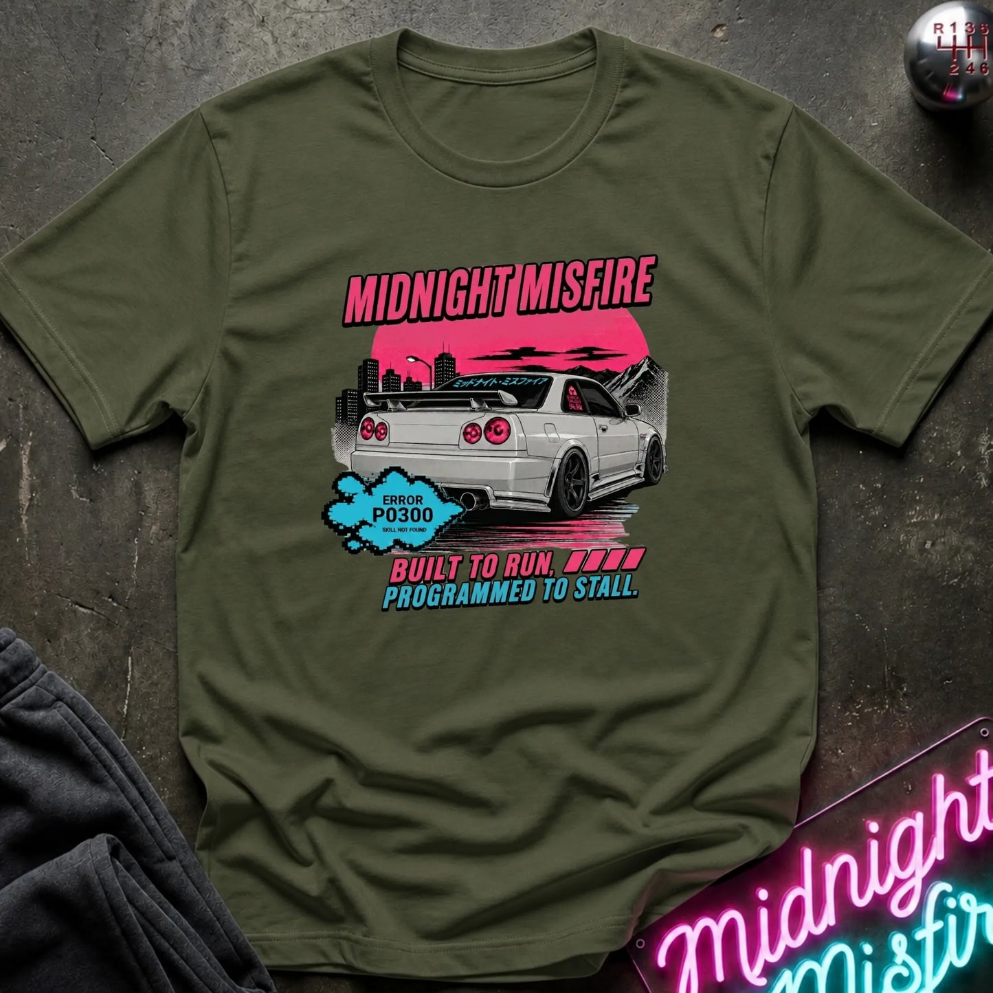 An olive green graphic t-shirt featuring a white Nissan Skyline R35 car with pink and blue text that reads 'MIDNIGHT MISFIRE' and 'BUILT TO RUN. PROGRAMMED TO STALL.