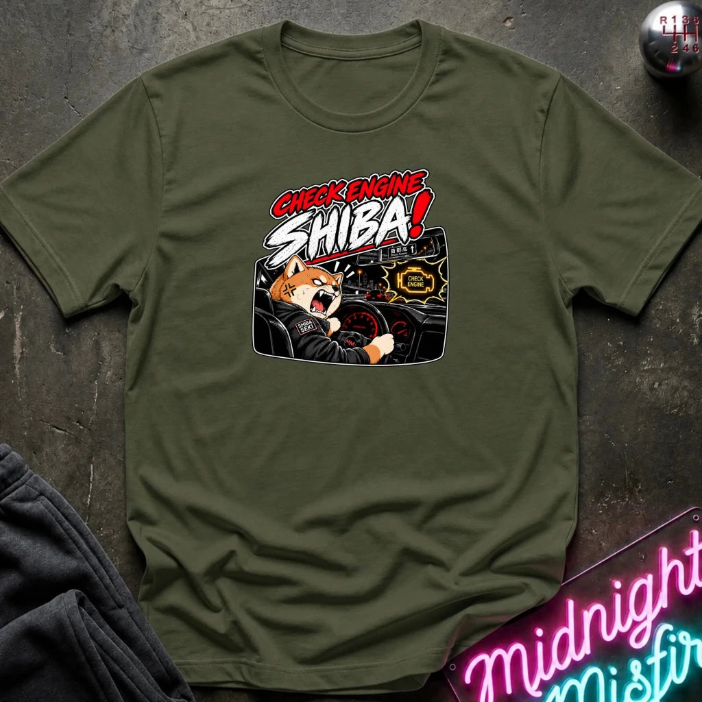 An olive green crewneck t-shirt featuring a graphic print of a Shiba Inu dog driving a car with the text 'CHECK ENGINE SHIBA!' in red and white, laid flat on a dark textured surface.