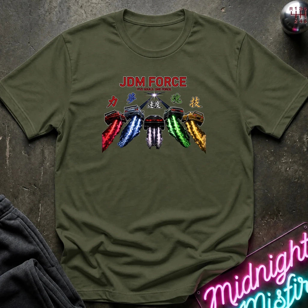An olive green short-sleeve t-shirt laid flat on a dark textured surface. The shirt features a graphic print with the text 'JDM FORCE' in red, followed by stylized Japanese characters and a row of colorful car headlights in red, blue, white, green, and gold.