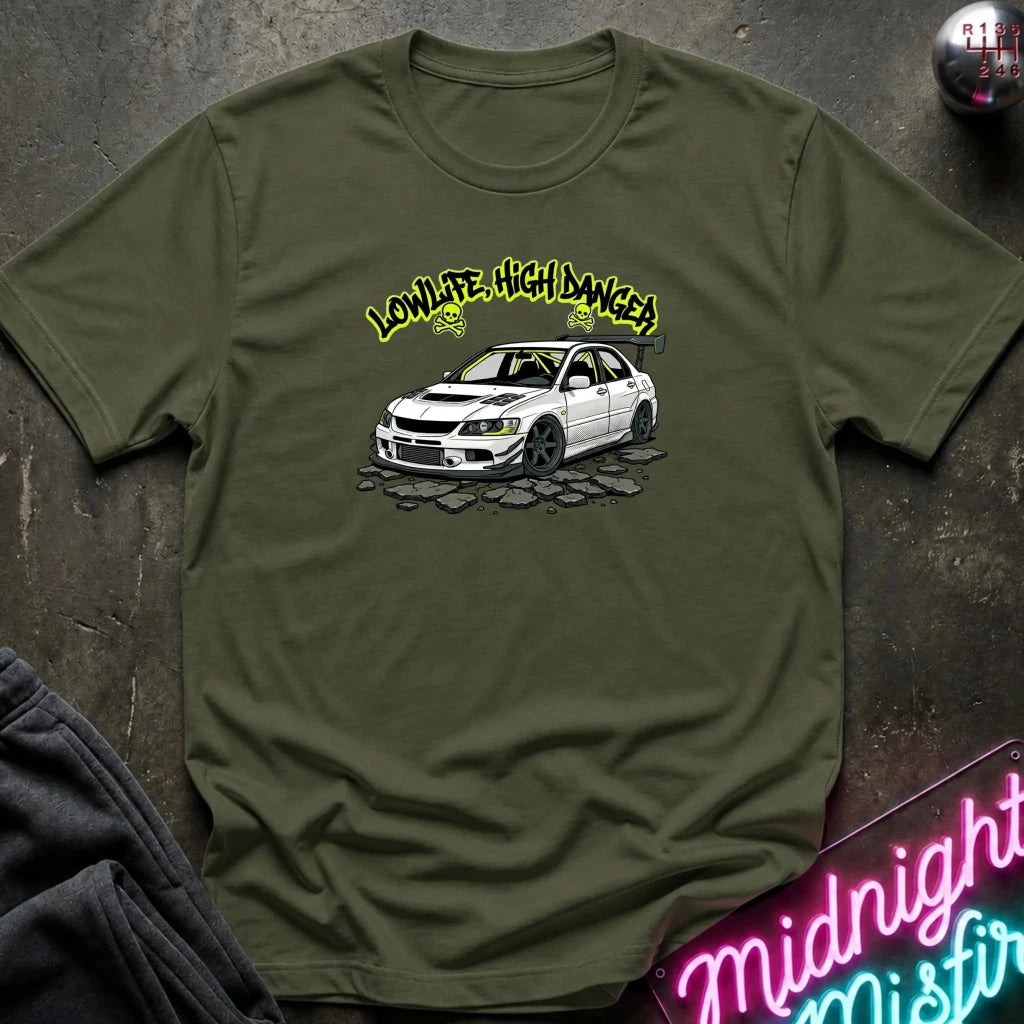 An olive green crewneck t-shirt laid flat on a dark textured surface. The shirt features a graphic print of a white sports sedan with yellow text reading 'LOWLIFE, HIGH DANGER!' above it.