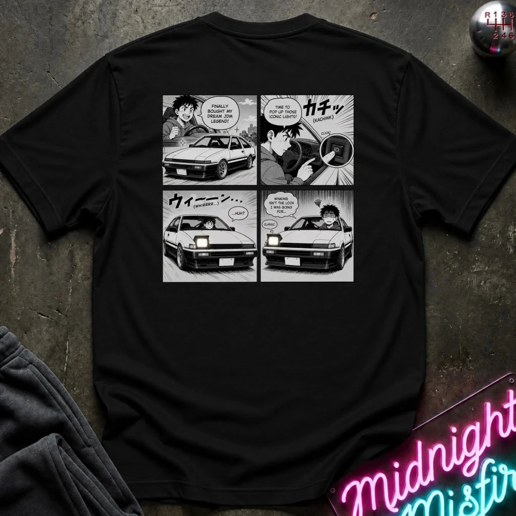 A black short-sleeve graphic t-shirt laid flat on a dark textured surface. The back of the shirt features a four-panel black and white manga-style comic print depicting characters and a car.
