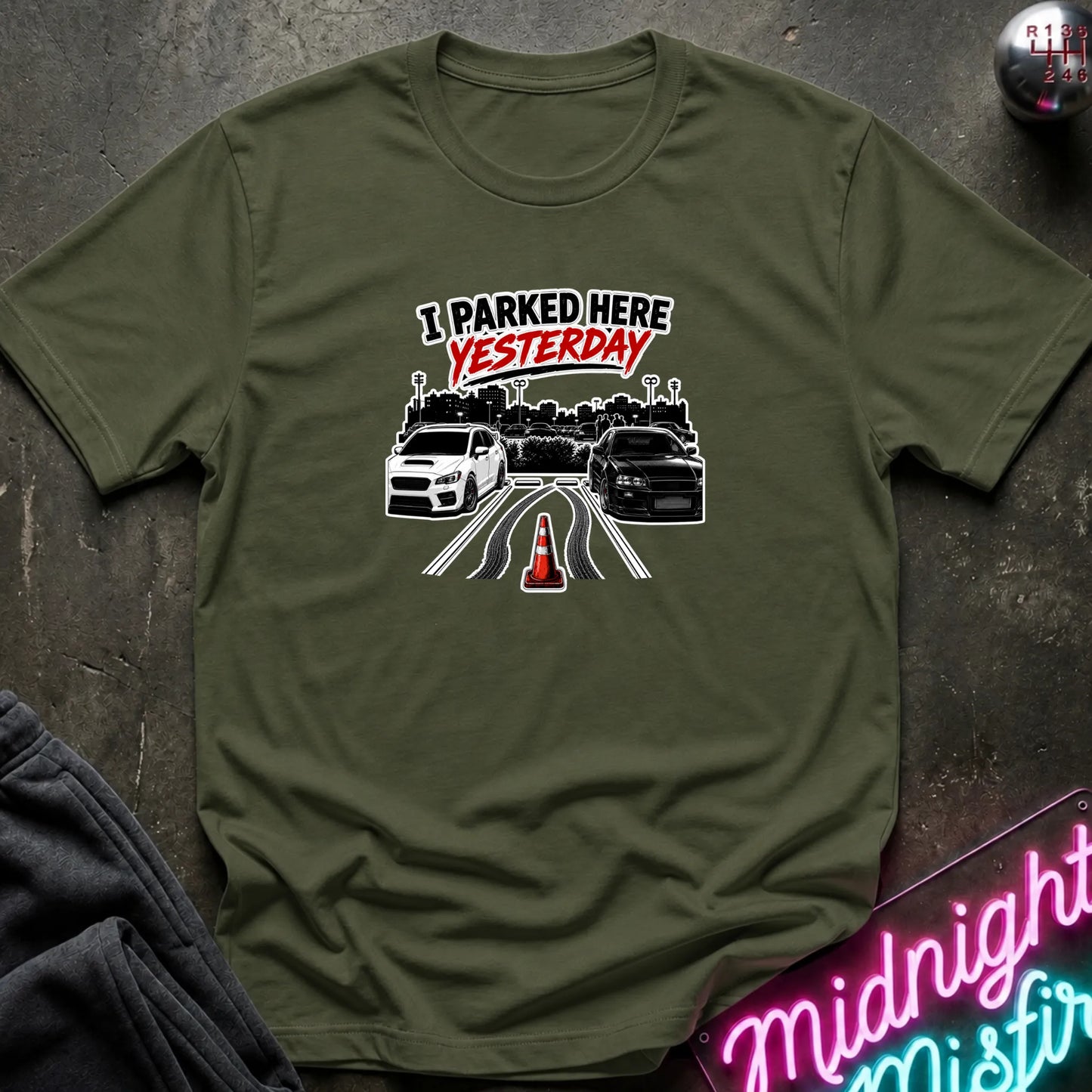 A olive green short-sleeved t-shirt  that has 2 cars parked in a space in between empty space with skid marks.