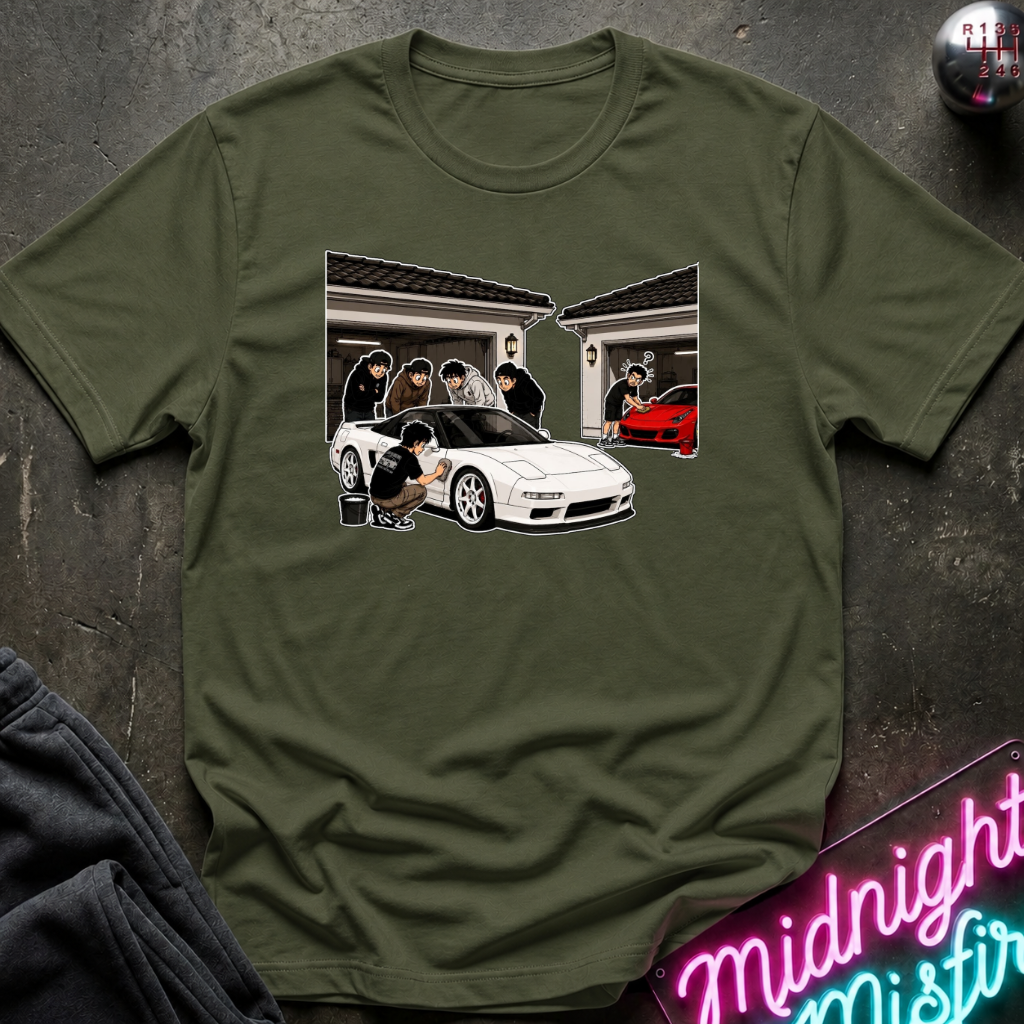 An olive green short-sleeve t-shirt with a graphic print of a white sports car and people in front of a house, styled flat on a dark textured background with a neon sign and dark pants partially visible.